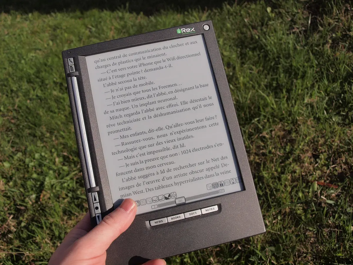 close-up of an anonymous hand holding a slim unbranded device showing a monochrome e-ink page, stylus beside it on a desk