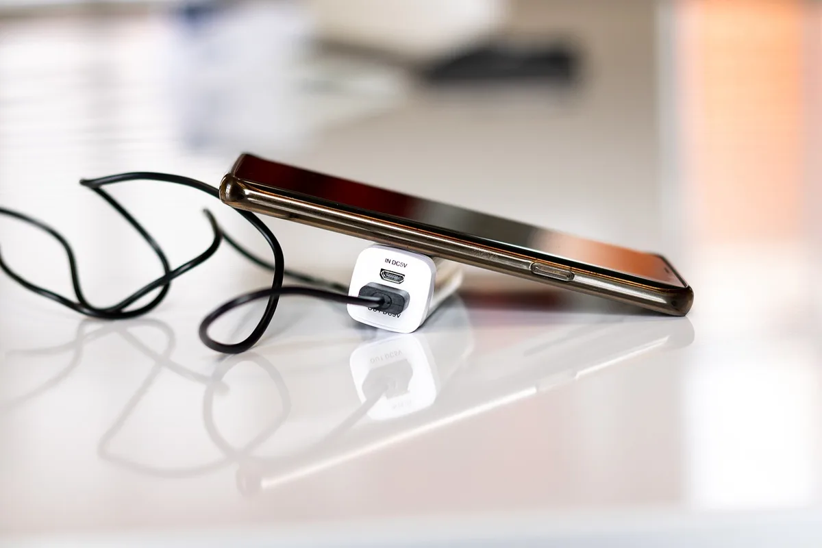 Wide shot of an anonymous unbranded smartphone lying on a table next to a notebook and charging cable, battery-focused composition