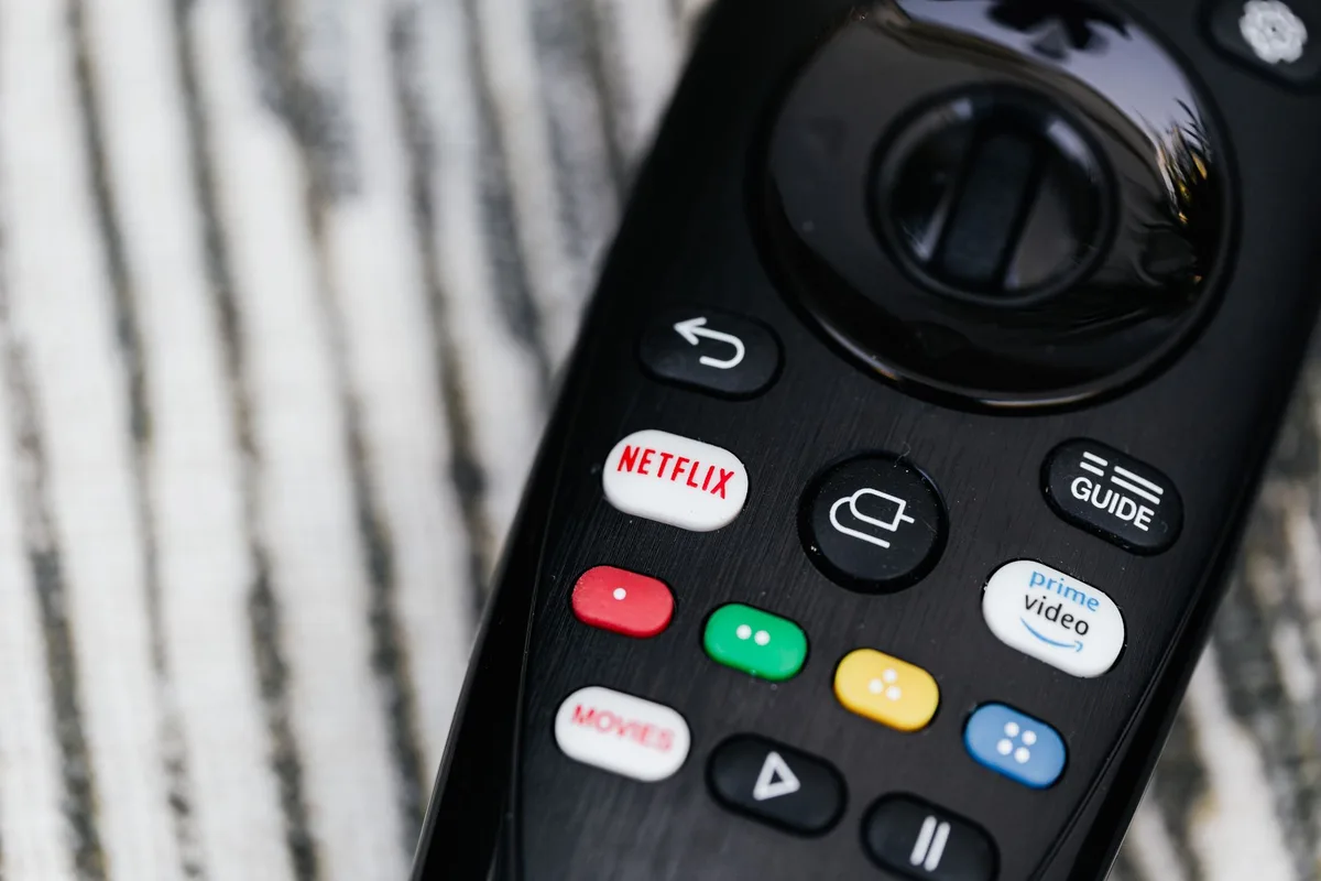 Close-up of an anonymous hand holding a slim unbranded remote control above a couch, soft-focus living room background, hint of a TV screen glow