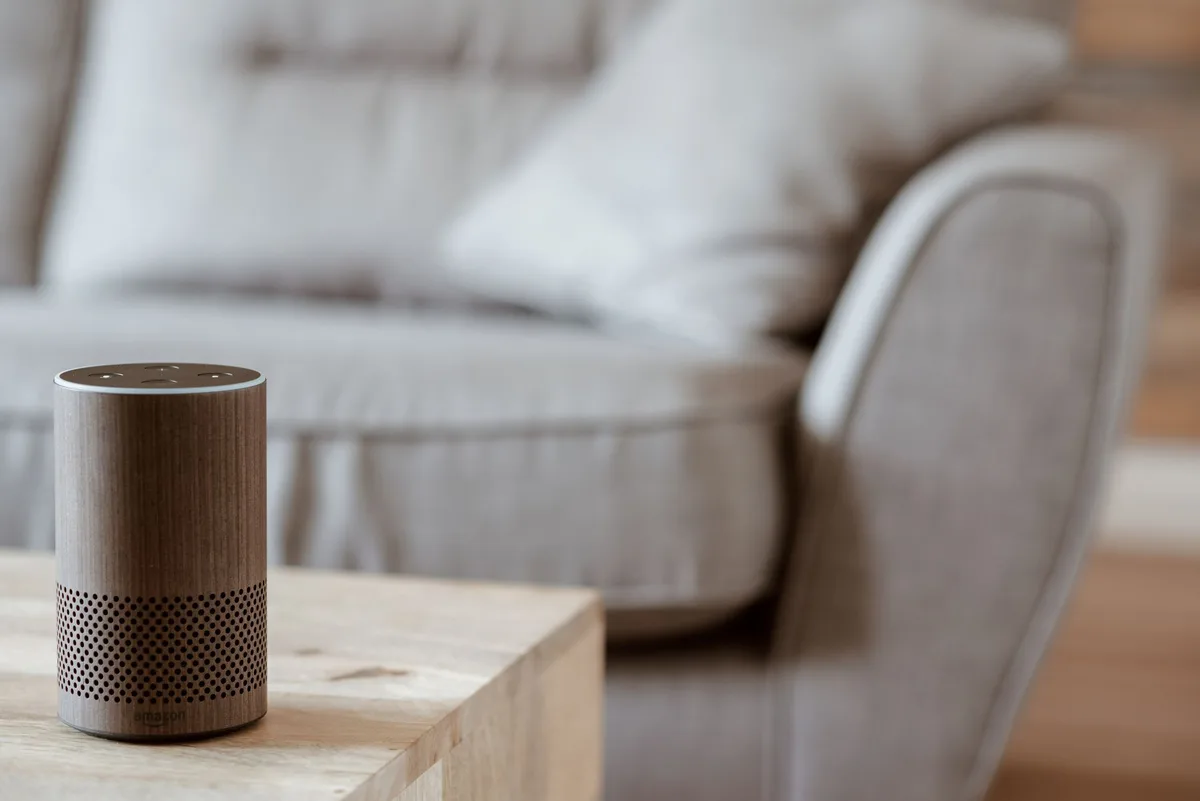A small smart speaker atop a nightstand casting soft colored light onto a pale wall, anonymous domestic scene suggesting ambient music and technology